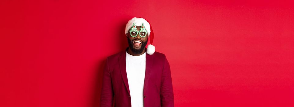Merry Christmas. Cheerful Black man wearing funny party glasses and santa hat, smiling joyful, celebrating winter holidays, standing over red background.