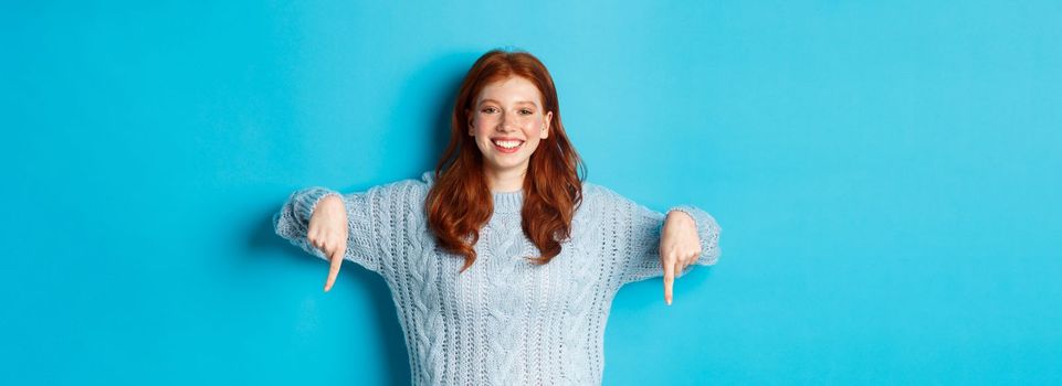 Winter holidays and people concept. Cheerful redhead woman in sweater pointing fingers down, smiling pleased at camera, showing promo, blue background.