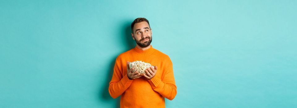 Handsome young man in orange sweater, looking thoughtful at upper left corner, holding popcorn, picking movie, blue background.