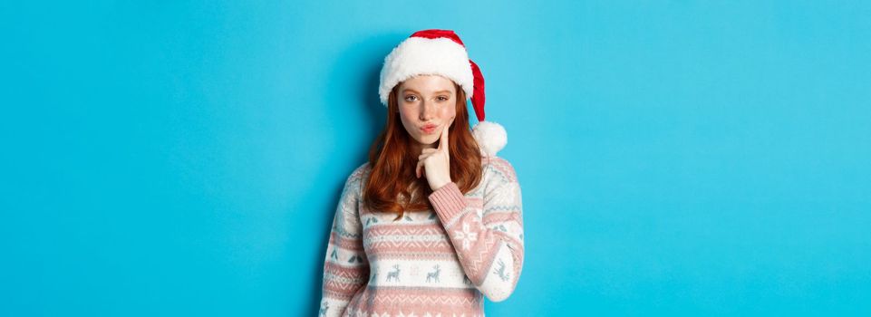Winter and Christmas Eve concept. Thoughtful redhead teen girl looking at camera, making choice or thinking, wearing santa hat, standing over blue background.