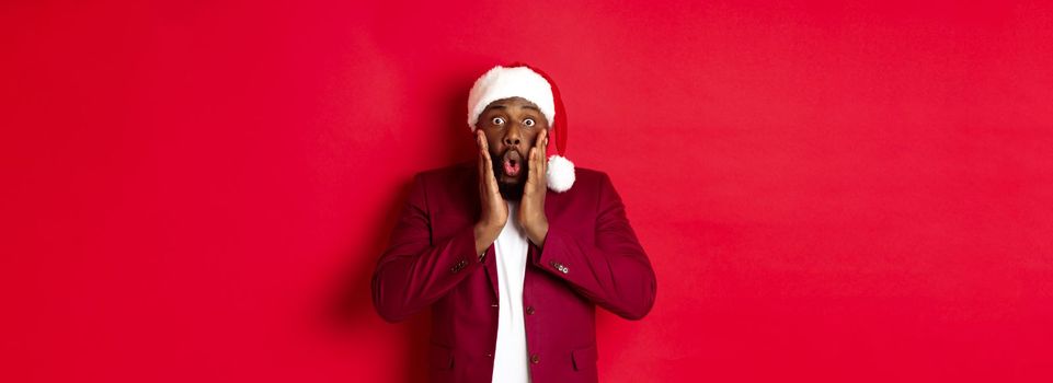 Christmas, party and holidays concept. Shocked Black man in santa hat reacting to new year offer, standing against red background.