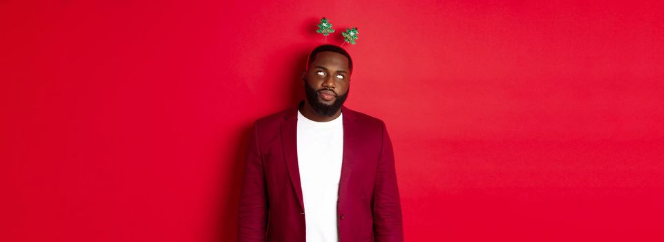 Merry Christmas. Annoyed african american man roll eyes, wearing silly headband on party, standing over red background.