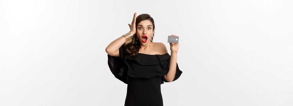 Fashion and shopping concept. Shocked woman in black dress showing credit card, looking in awe at camera, white background.