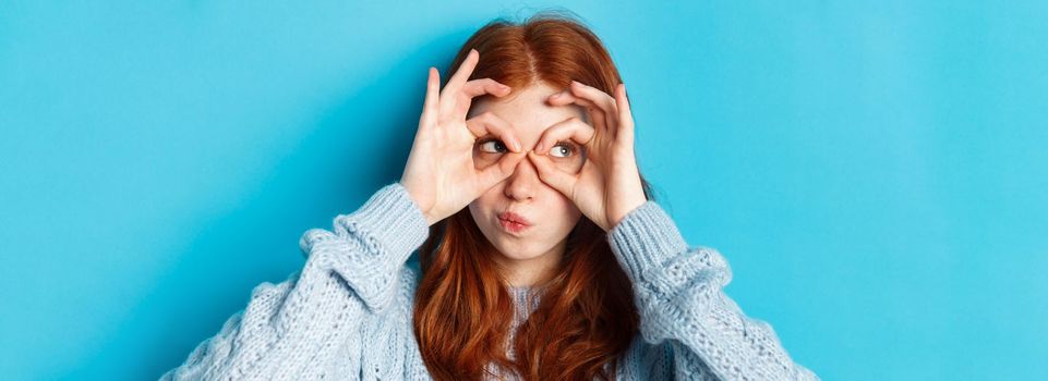 Headshot of funny redhead girl thinking, looking through finger glasses with thoughtful face, standing against blue background.