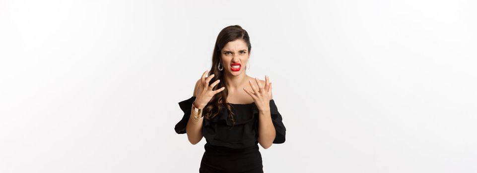 Fashion and beauty. Angry woman in black dress, shouting mad and shaking hands, grimacing outraged at camera, standing over white background.