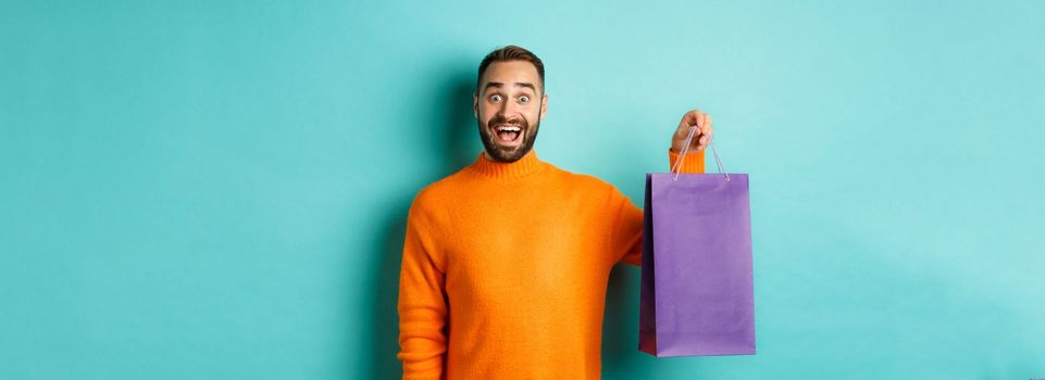 Excited adult man holding purple shopping bag and smiling, going to mall, standing over turquoise background.
