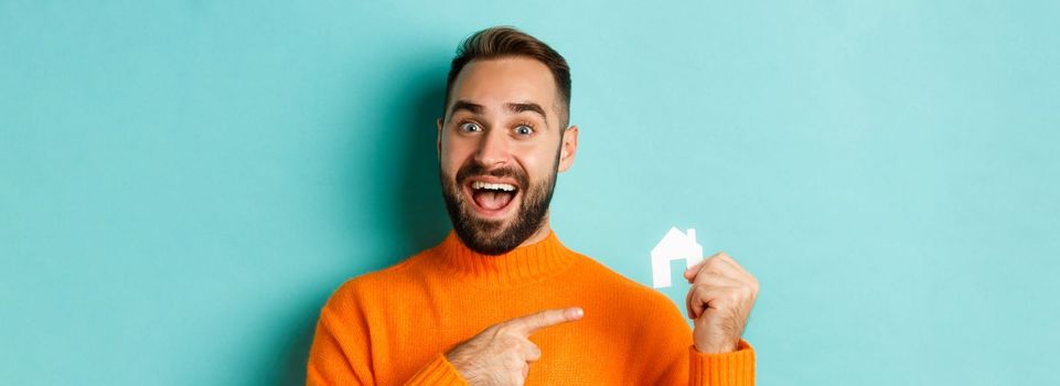 Real estate. Excited bearded man pointing at small paper house maket, standing amused over light blue background.
