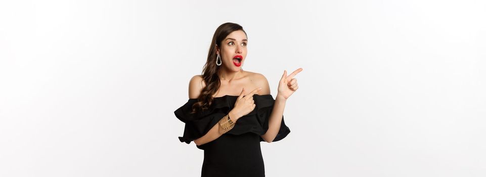 Fashion and beauty. Amazed beautiful woman in black dress open mouth fascinated, pointing fingers and looking left, white background.