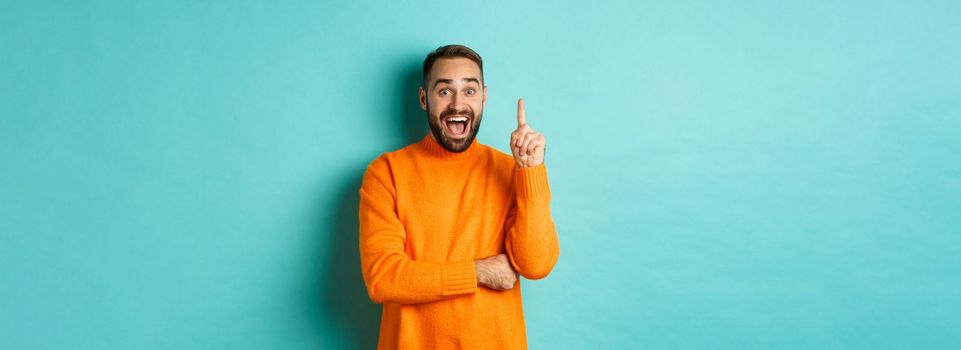 Handsome caucasian man having an idea, raising finger up and saying his plan, standing excited against turquoise background.