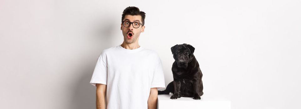 Image of dog owner and cute black pug looking at camera surprised and amazed, standing over white background.