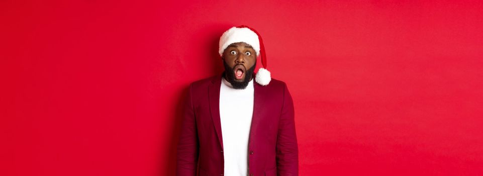 Christmas, party and holidays concept. Shocked Black man in santa hat drop jaw and stare at camera, stand against red background.