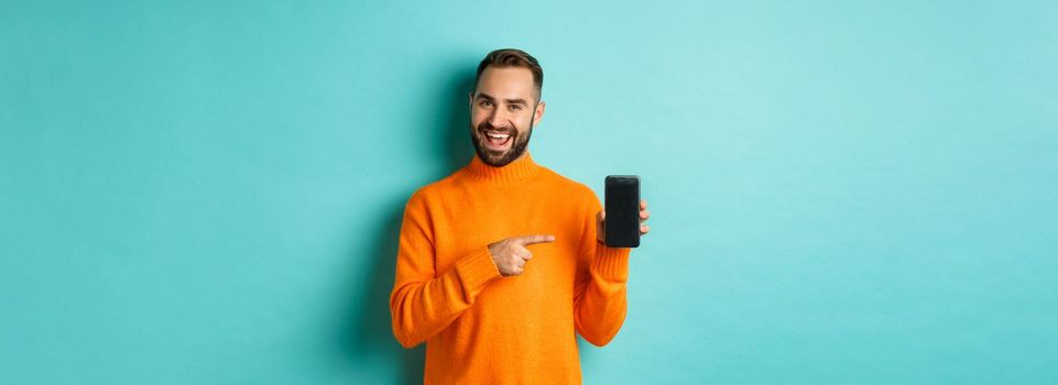 Handsome bearded man in orange sweater, pointing finger at mobile phone screen, showing application smartphone, standing over light blue background.