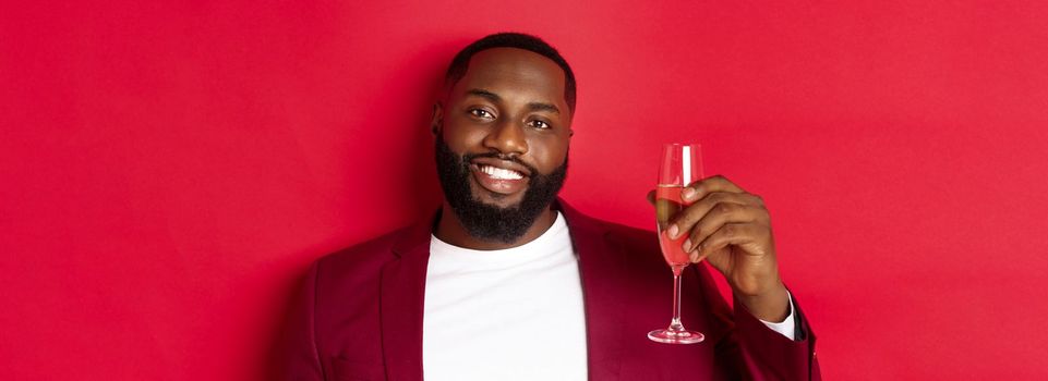 Close-up of handsome smiling Black man saying toast, raising glass of champagne for merry christmas and happy new year, celebrating against red background.