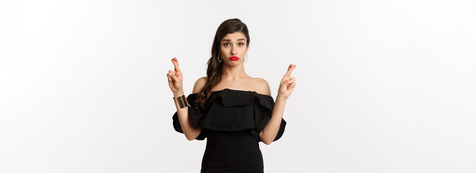 Fashion and beauty. Hopeful silly woman in black dress making wish, holding fingers crossed for good luck, standing over white background.