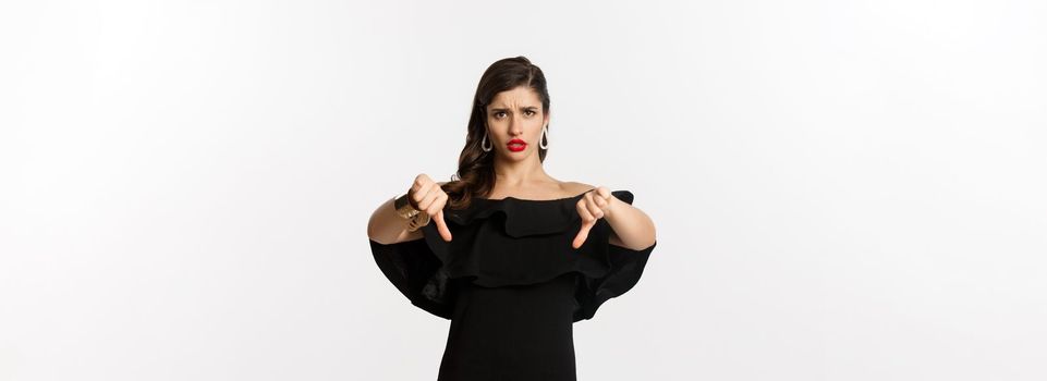 Fashion and beauty. Disappointed and upset woman in black dress, showing thumbs down, dislike something bad, judging over white background.