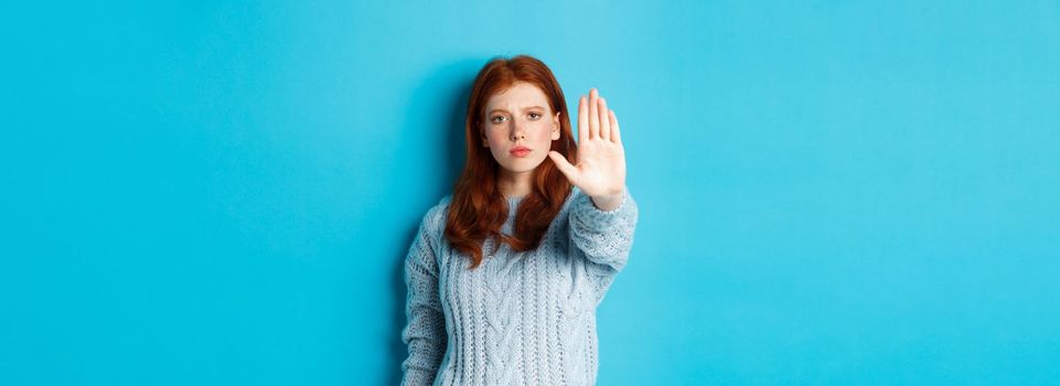 Serious and confident redhead girl telling to stop, saying no, showing extended palm to prohibit action, standing over blue background.