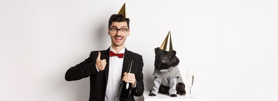 Small black dog wearing party hat and standing near happy man celebrating holiday, owner showing thumb-up and holding champagne bottle, white background.