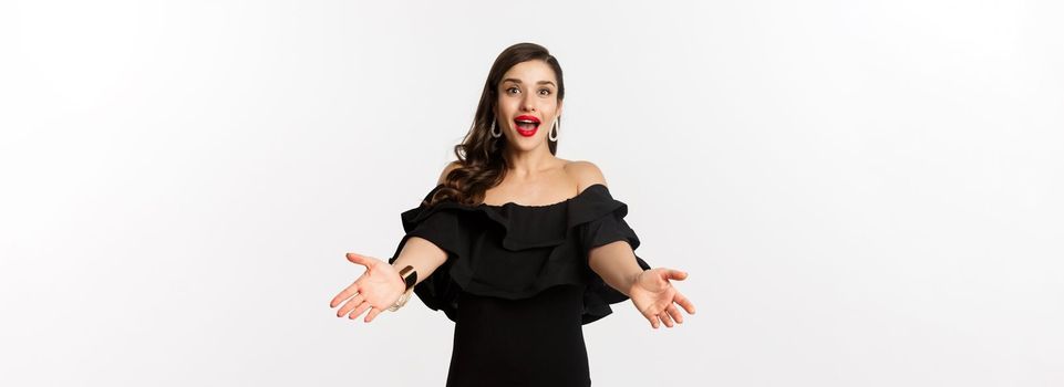 Fashion and beauty concept. Happy young woman in glamour black dress, reaching hands forward to take something, waiting for hug, white background.