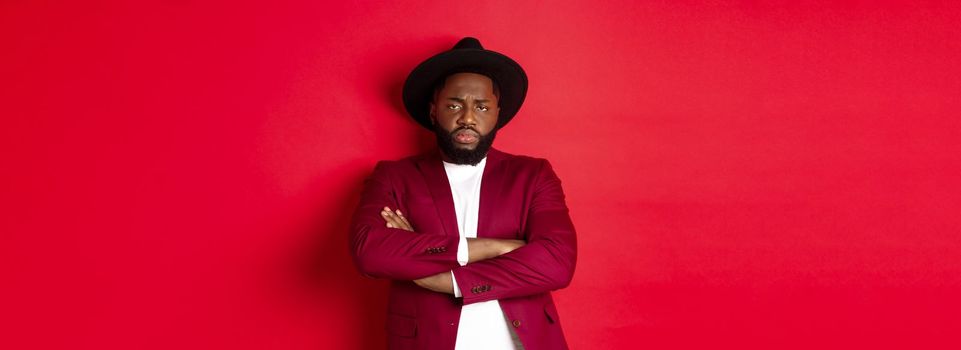 Reluctant and upset Black man waiting for apology, cross arms on chest and looking offended at camera, red background. Copy space