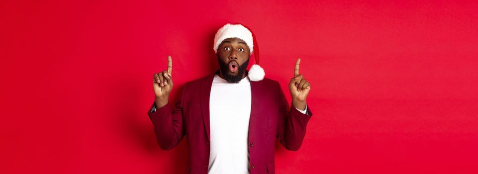 Christmas, party and holidays concept. Impressed Black man showing offer, pointing fingers up at logo, saying wow, standing against red background.