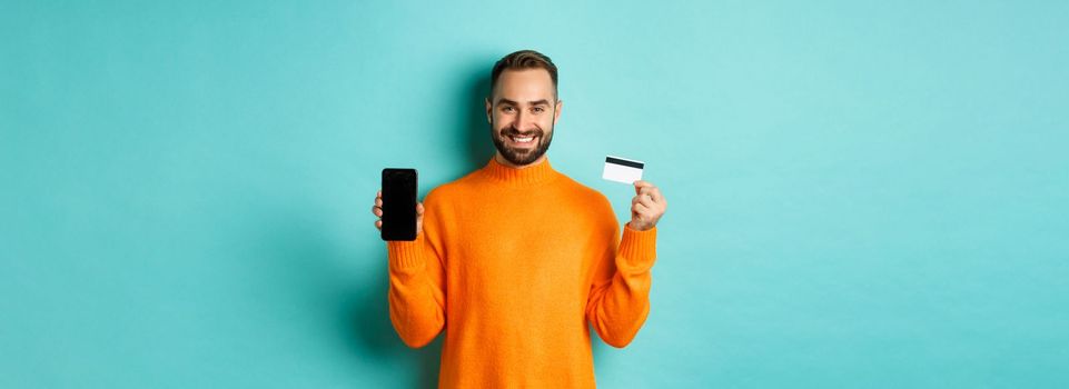 Online shopping. Happy attractive guy showing mobile phone screen and credit card, smiling satisfied, standing over light blue background.
