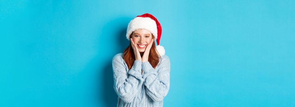 Winter holidays and Christmas Eve concept. Cheerful redhead girl in santa hat, celebrating New Year, blushing and smiling happy, standing over blue background.