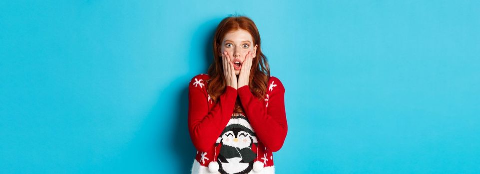 Winter holidays and Christmas Eve concept. Impressed and speechless redhead girl gasping, staring at camera with disbelief, standing in xmas sweater against blue background.
