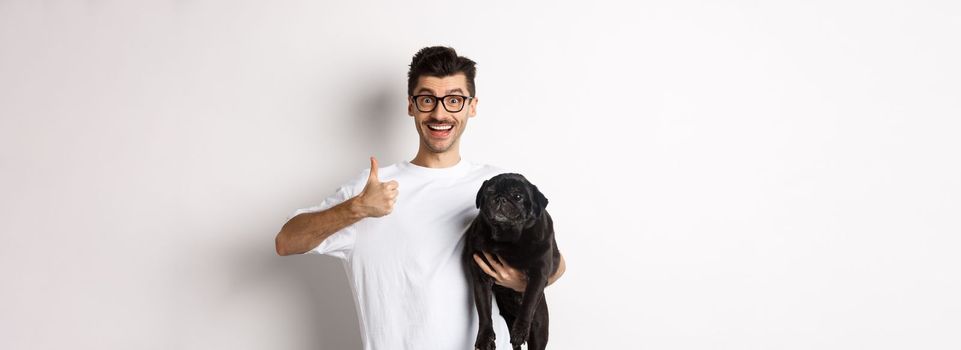 Cheerful handsome man holding dog and showing okay sign, approve or recommend product. Hipster guy carry cute black pug and looking satisfied, white background.