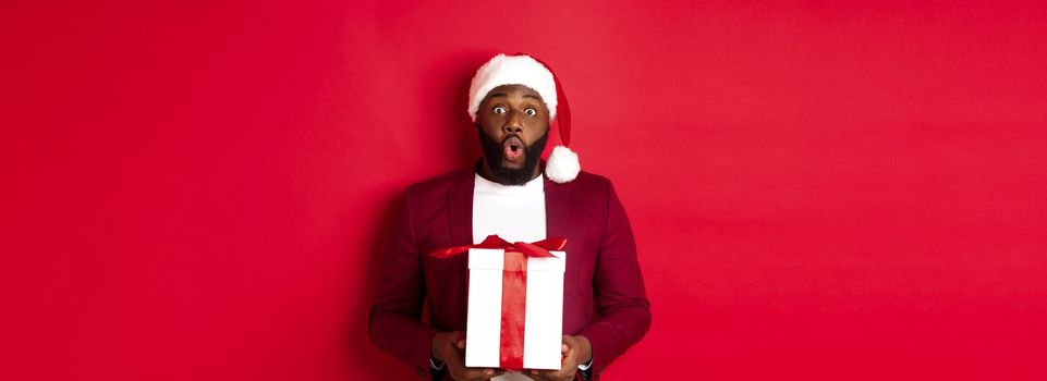 Christmas, New Year and shopping concept. Surprised african american man in santa hat saying wow, holding holiday gift and looking at camera amazed, red background.