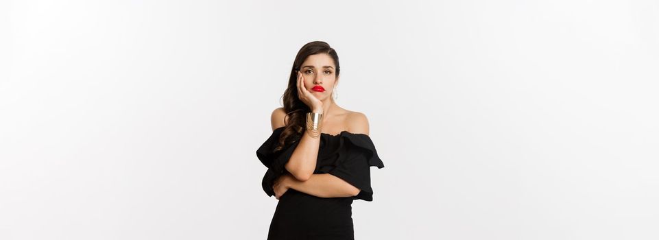 Fashion and beauty. Bored stylish woman in makeup, black dress, waiting for something, looking sad and gloomy, standing against white background.