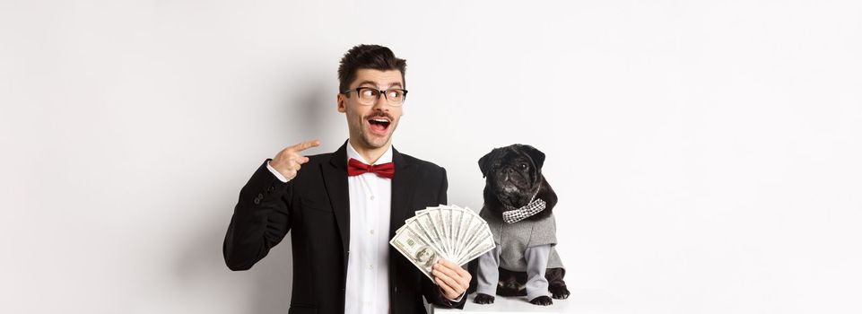 Happy young man in suit earn money with his dog. Guy rejoicing, holding dollars and pointing left, black pug in costume staring at camera, white background.