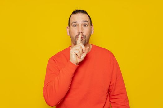 A serious handsome man in a red sweater, puts his index finger to his lips, tries to keep the conspiracy, looks to the side and says: Shh, be quiet please. Isolated shot of man shows silence sign