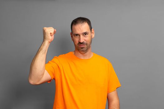 Bearded hispanic man wearing an orange t-shirt celebrating something with his fist up isolated on gray studio background