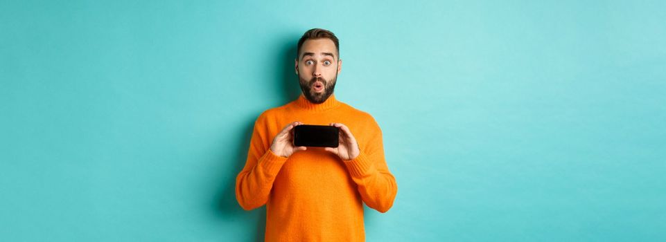 Impressed man showing smartphone screen, stare at camera amazed, demonstrate display, standing over light blue background.