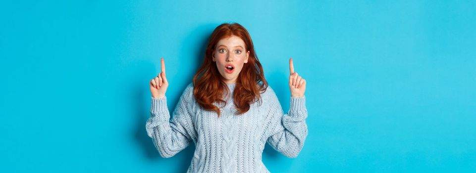 Winter holidays and people concept. Amazed redhead teenage girl pointing fingers up, showing advertisement and staring impressed at camera, blue background.