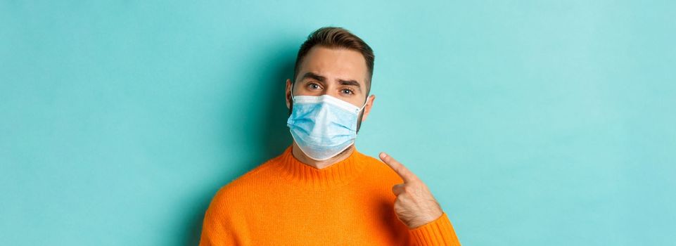 Covid-19, social distancing and quarantine concept. Upset man in complaining on face mask, pointing at it and looking bothered, standing over light blue background.
