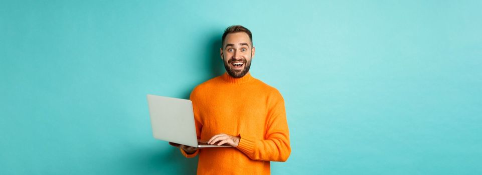Excited smiling man working on laptop, staring at camera happy, standing in orange sweater against turquoise background.