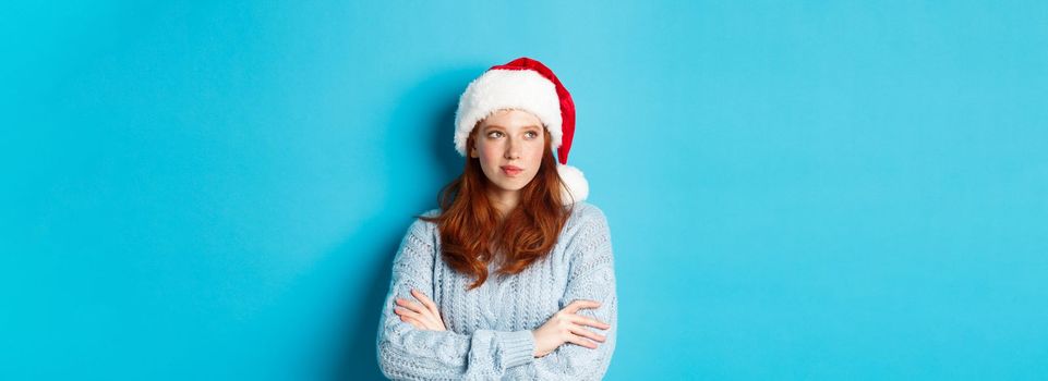 Winter holidays and Christmas Eve concept. Thoughtful redhead woman in Santa hat and sweater, looking left and pondering, making xmas plans, standing over blue background.