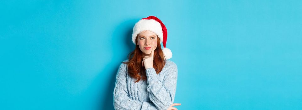 Winter holidays and Christmas Eve concept. Silly redhead girl with freckles, wearing santa hat and thinking, planning New Year celebration, standing over blue background.