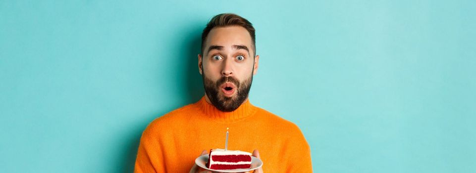 Close-up of happy adult man celebrating birthday, holding bday cake with candle and making wish, standing against turquoise background.