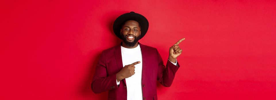 Winter holidays and shopping concept. Happy Black man pointing left and smiling, showing new year promo offer on red background.