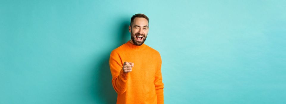 Cheeky attractive man congrats you, praising good job, winking and pointing at camera, standing over light blue background.