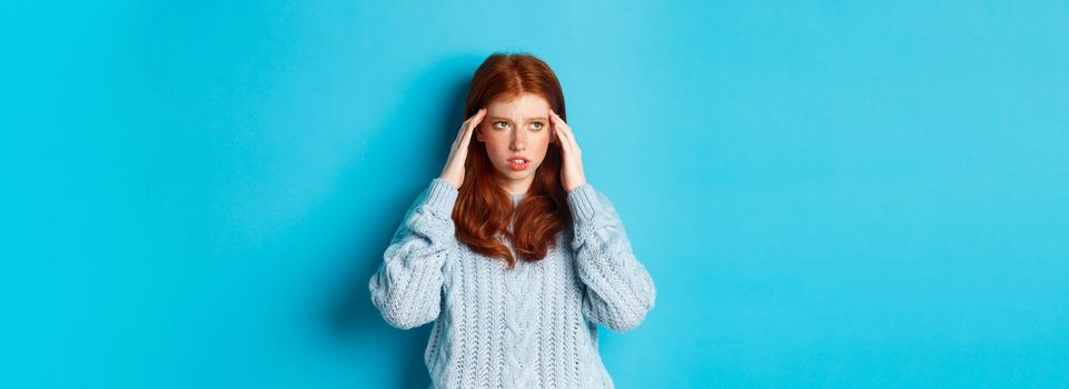 Young tensed redhead college girl trying to think, rubbing temples and looking up, thinking, standing over blue background.