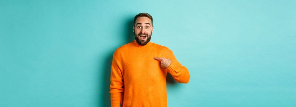 Excited man pointing at himself, looking amazed and happy, being chosen, standing over light blue background.
