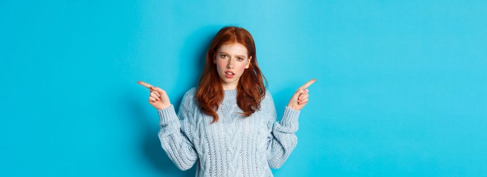 Confused redhead girl in sweater pointing fingers sideways, staring at camera doubtful, standing against blue background. Copy space