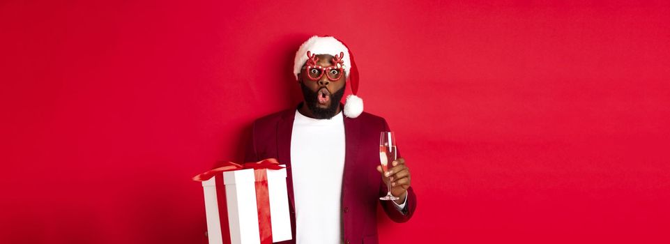 Christmas. Handsome african american man in party glasses and santa hat, holding new year gift and glass of champagne, wishing happy holidays, red background.