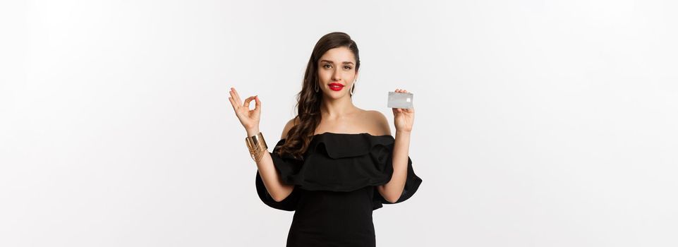 Beauty and shopping concept. Gorgeous woman in luxury jewelry and black dress, showing okay sign and credit card, standing over white background.