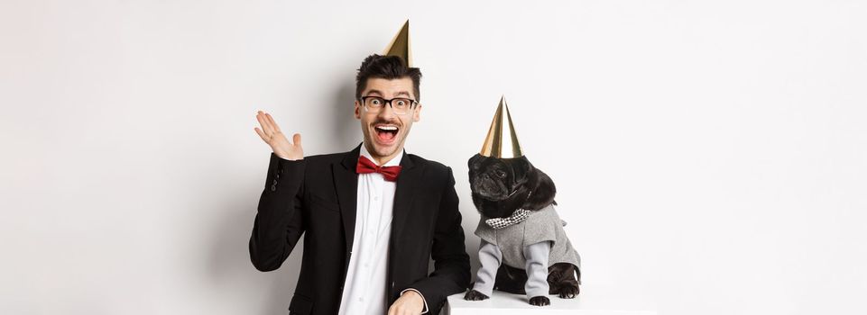 Happy young man and cute black dog wearing party cones, celebrating birthday, guy friendly saying hello and waving hand, standing over white background.