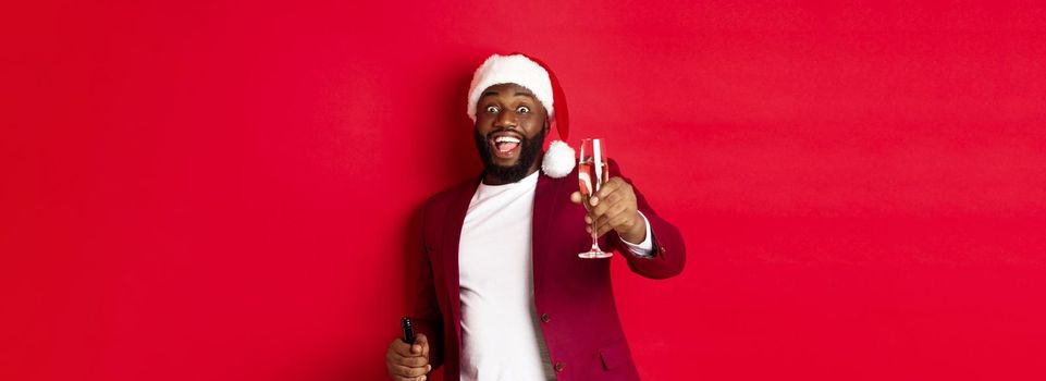 Christmas, party and holidays concept. Handsome Black man in santa hat raising glass of champagne and smiling, saying toast, celebrating New Year, red background.