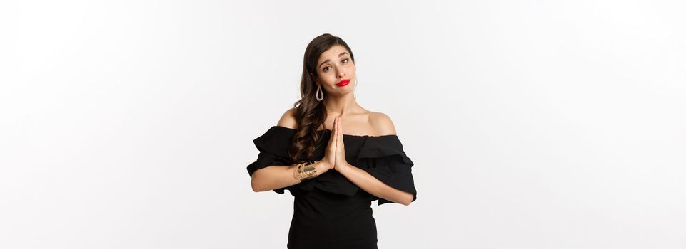 Fashion and beauty. Pretty stylish woman asking for help, begging for something, say please with hands pressed together, standing in black dress, white background.